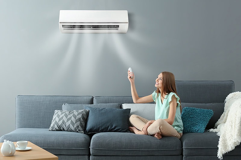 Young woman switching on air conditioner while sitting on sofa at home.